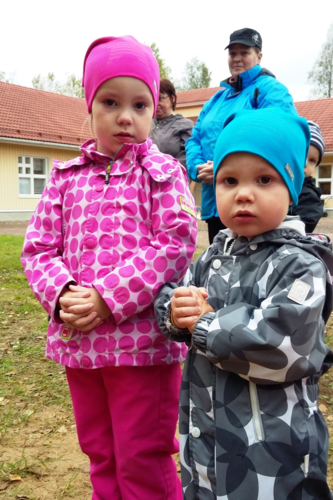 Kaksi lasta seisoo ulkona ja pitävät kädet ristissä. Taustalla kaksi aikuista.